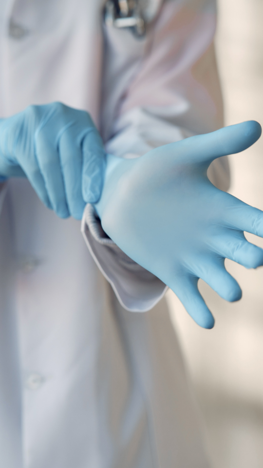 En bild som visar person i vit rock sätta på sig blåa gummi handskar